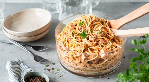 Spaghettisalat in einer Glasschüssel, zwei Hände halten ein Salatbesteck mit Salat –   Foto: Anna Gieseler