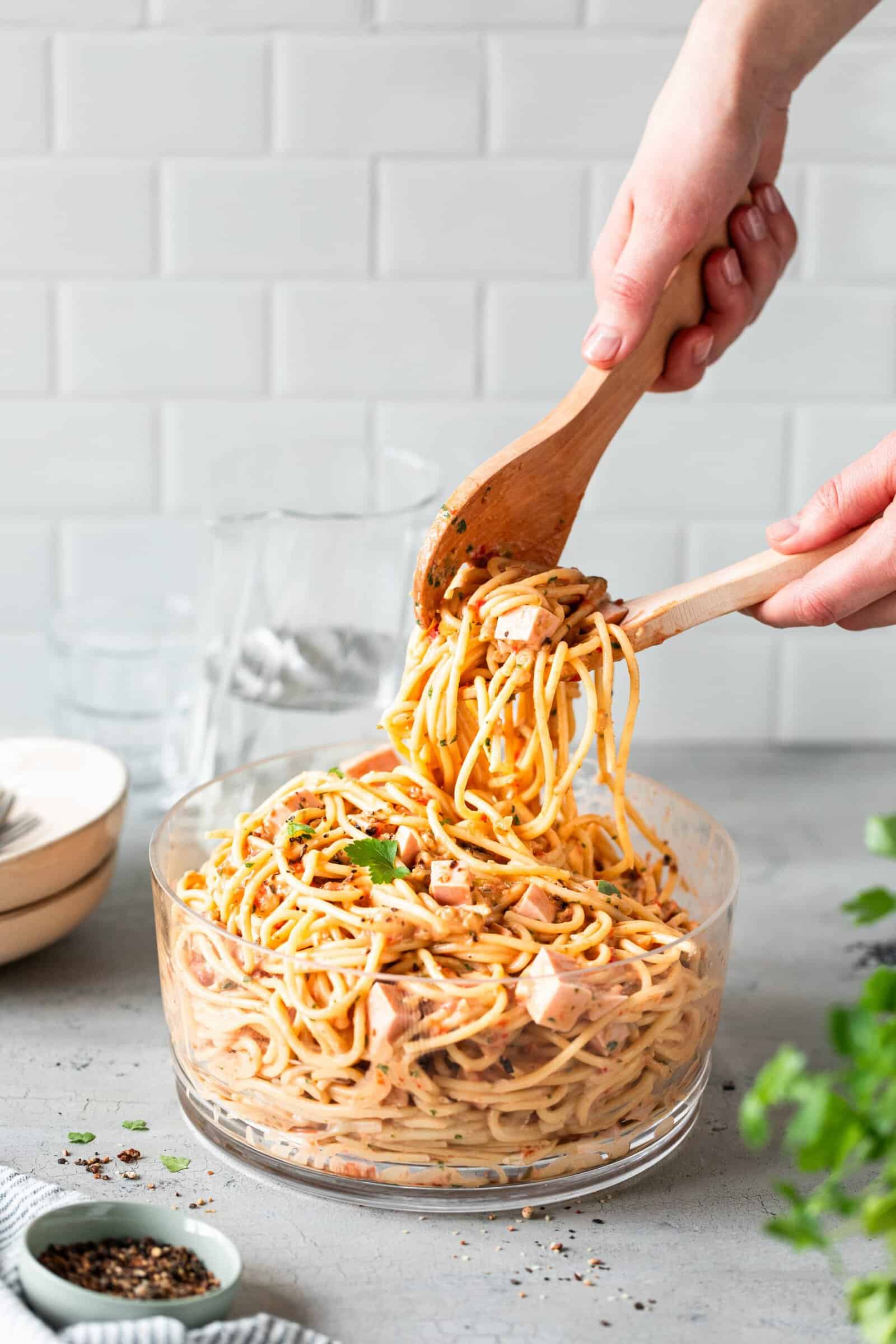 Spaghettisalat in einer Glasschüssel, zwei Hände halten ein Salatbesteck mit Salat –   Foto: Anna Gieseler